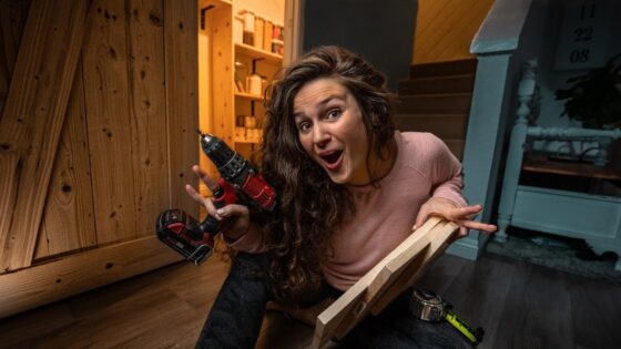 Woman kneeling on a workshop floor holds a cordless drill and a wooden plank with a surprised expression, surrounded by tools and wood.
