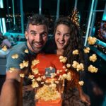 Smiling couple takes a selfie with a red popcorn machine popping popcorn around them in a blue-lit living room.
