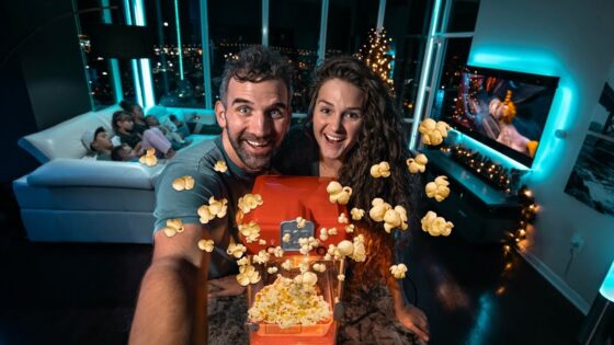 Smiling couple takes a selfie with a red popcorn machine popping popcorn around them in a blue-lit living room.