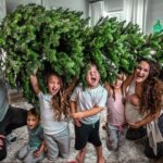 Family of six posing in a living room under a large evergreen Christmas tree, all smiling and excited.