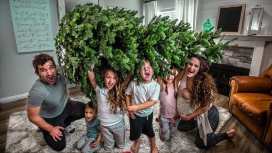 Family of six posing in a living room under a large evergreen Christmas tree, all smiling and excited.