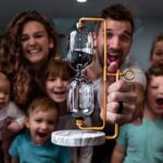 Family gathered around a man proudly holding a copper-framed hourglass device for a science demo in a cozy living room beyond.