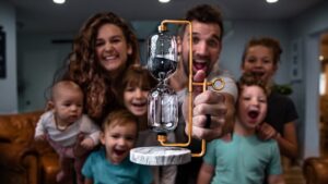 Family gathered around a man proudly holding a copper-framed hourglass device for a science demo in a cozy living room beyond.