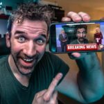 Man with a big smile holds a smartphone showing a breaking news broadcast with anchors on screen, in a home studio setting.