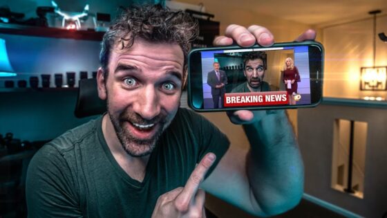 Man with a big smile holds a smartphone showing a breaking news broadcast with anchors on screen, in a home studio setting.