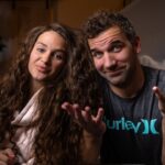 A smiling couple sits close together in a cozy living room, looking at the camera with relaxed expressions.