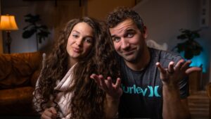 A smiling couple sits close together in a cozy living room, looking at the camera with relaxed expressions.
