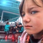 Close-up of a young girl with braided hair and blue eyes, looking at the camera, with a blurred cafe scene in the background under a teal canopy.