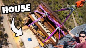 Piano strapped with purple lifting straps being hoisted from a rooftop, with a man shouting from a balcony wearing sunglasses.