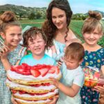 A woman with children in a strawberry field, proudly holding a large strawberry-topped layered cake.