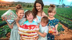 A woman with children in a strawberry field, proudly holding a large strawberry-topped layered cake.
