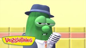 Green cucumber character in a hat and suit sings into a vintage microphone on a stage, with the VeggieTales logo in the corner.