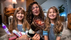 Woman and two girls smile at the camera while holding a hair dryer and several spray bottles in a cozy living room setting.