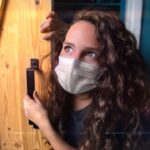 Young woman wearing a face mask peers around a wooden door, gripping the handle and looking upward.