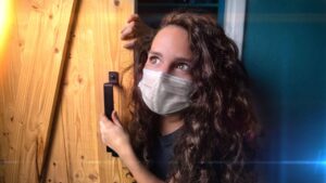 Young woman wearing a face mask peers around a wooden door, gripping the handle and looking upward.