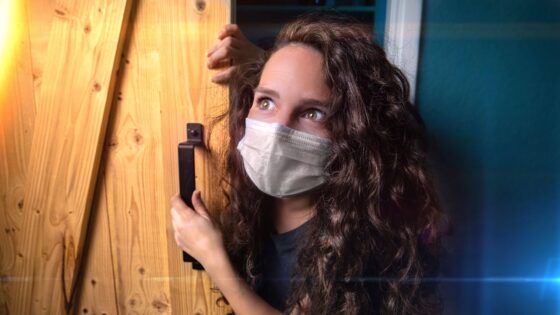 Young woman wearing a face mask peers around a wooden door, gripping the handle and looking upward.