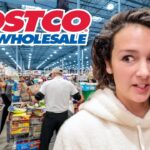 Woman with long curly hair in a white hoodie stands in a busy Costco store, shopping carts and shelves in the background.