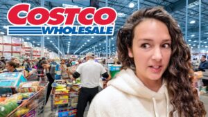 Woman with long curly hair in a white hoodie stands in a busy Costco store, shopping carts and shelves in the background.