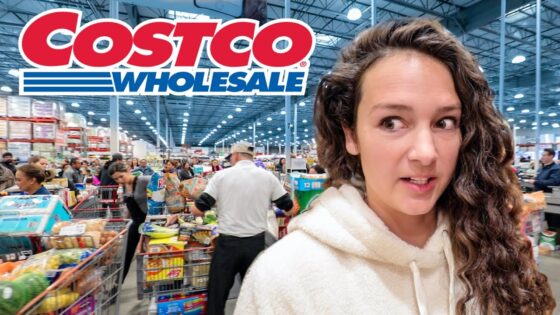 Woman with long curly hair in a white hoodie stands in a busy Costco store, shopping carts and shelves in the background.