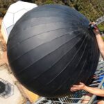 Man in a blue shirt and sunglasses hangs from a safety railing on a tall tower, gripping a large black spherical object with seams; a white geodesic dome is visible nearby.