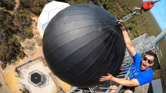 Man in a blue shirt and sunglasses hangs from a safety railing on a tall tower, gripping a large black spherical object with seams; a white geodesic dome is visible nearby.