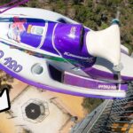 Thrill ride scene: a purple Kawasaki jet ski is mounted above a platform while a man in blue stands on the railing, giving a thumbs-up.