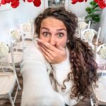 Woman takes a selfie in a bright room with red petal decorations above and white chairs with floral accents in the background.