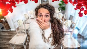 Woman takes a selfie in a bright room with red petal decorations above and white chairs with floral accents in the background.