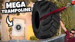 Thumbnail of a man in sunglasses on a high railing, gripping a giant tire attached to cords for a 'mega trampoline' stunt, with a hexagonal landing pad on the ground marked by a question mark and a bold arrow.