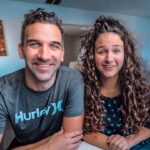 Smiling man and woman sit at a table in a bright, modern room with world map wall art behind them.