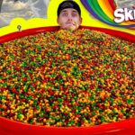 Man's head sticking out from a giant red tub filled with multicolored Skittles candy, with a bright yellow border and Skittles logo in the background.