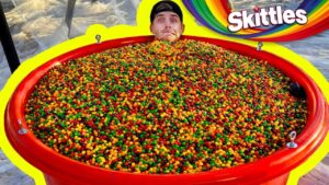Man's head sticking out from a giant red tub filled with multicolored Skittles candy, with a bright yellow border and Skittles logo in the background.