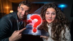 Two people smile at the camera while presenting a laptop with a large red question mark on screen.