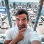 Man taking a selfie on a glass-walled observation deck with a city skyline below