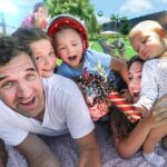 Family selfie on a sunny day at the park with kids, a baby wearing a helmet, and a red balloon nearby.