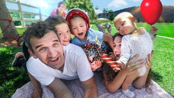 Family selfie on a sunny day at the park with kids, a baby wearing a helmet, and a red balloon nearby.