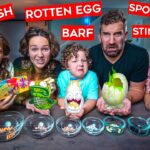 Family of five posing with gross-food challenge props labeled Dead Fish, Rotten Egg, Spoiled Milk, Barf, and Stink Bug; bowls of jelly beans in front.