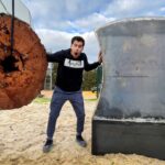 Man in a black hoodie with a surprised expression stands between a huge tree trunk slice and a large curved metal sculpture outdoors, touching both.