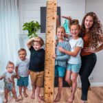 A woman and six children pose indoors with a tall wooden ruler showing height measurements, as they smile for the photo.