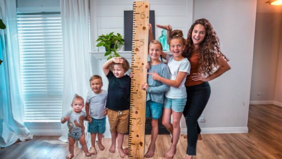 A woman and six children pose indoors with a tall wooden ruler showing height measurements, as they smile for the photo.