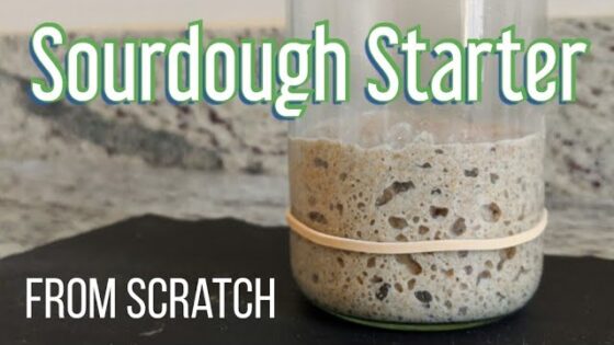 Jar of bubbly sourdough starter with a rubber band, labeled 'Sourdough Starter From Scratch' on a dark surface