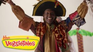 Smiling performer in a colorful pirate-like costume raises arms, VeggieTales logo visible in the corner.