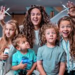 A woman with long curly hair holds up large scissors behind four children who are posing and smiling in a cozy living room, as if about to give a playful haircut.