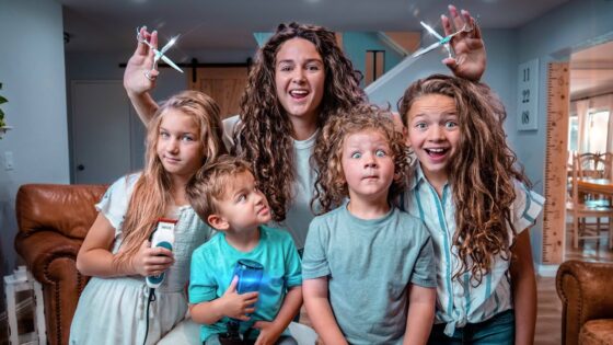 A woman with long curly hair holds up large scissors behind four children who are posing and smiling in a cozy living room, as if about to give a playful haircut.