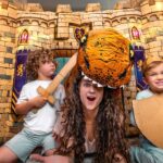 Mother wearing a tiger headpiece plays with two kids inside a large cardboard castle, one child holding a wooden sword and the other a shield, on a living room floor.