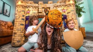 Mother wearing a tiger headpiece plays with two kids inside a large cardboard castle, one child holding a wooden sword and the other a shield, on a living room floor.