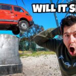 Man in a green sweatshirt with a shocked expression as a red car is hoisted by a crane above a concrete pedestal against a blue sky.