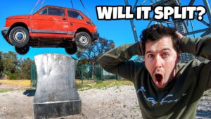Man in a green sweatshirt with a shocked expression as a red car is hoisted by a crane above a concrete pedestal against a blue sky.