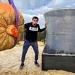 Man with a surprised expression poses beside a gigantic pumpkin hanging by purple straps next to a large curved metal sculpture in a sandy outdoor area.