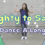 Young woman in a blue dress dances with arms raised in front of a gray brick wall, with colorful mountains and the title 'Mighty to Save Dance-A-Long' on the sides.